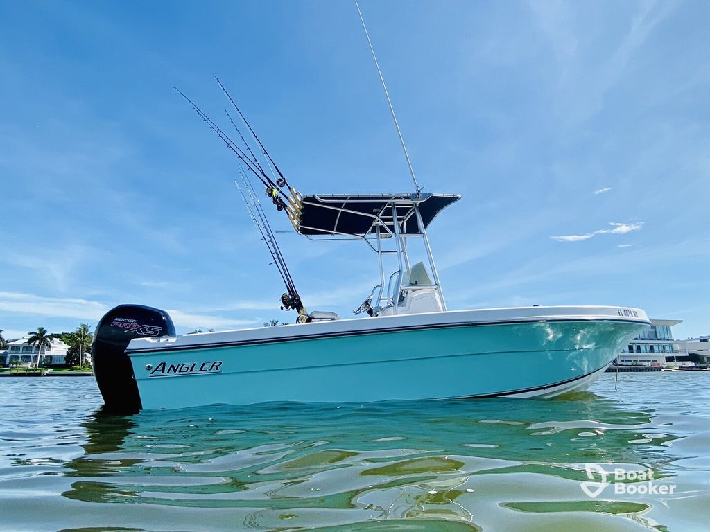 A light blue Angler fishing boat with a black outboard motor and fishing rods floats on calm water near shoreline buildings under a clear sky.