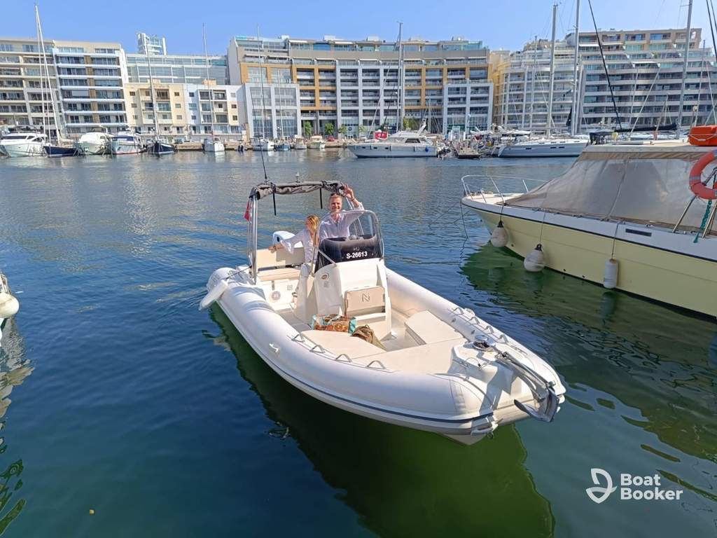 A motorboat with two people aboard's docked in a marina, with modern apartment buildings and other boats in the background.