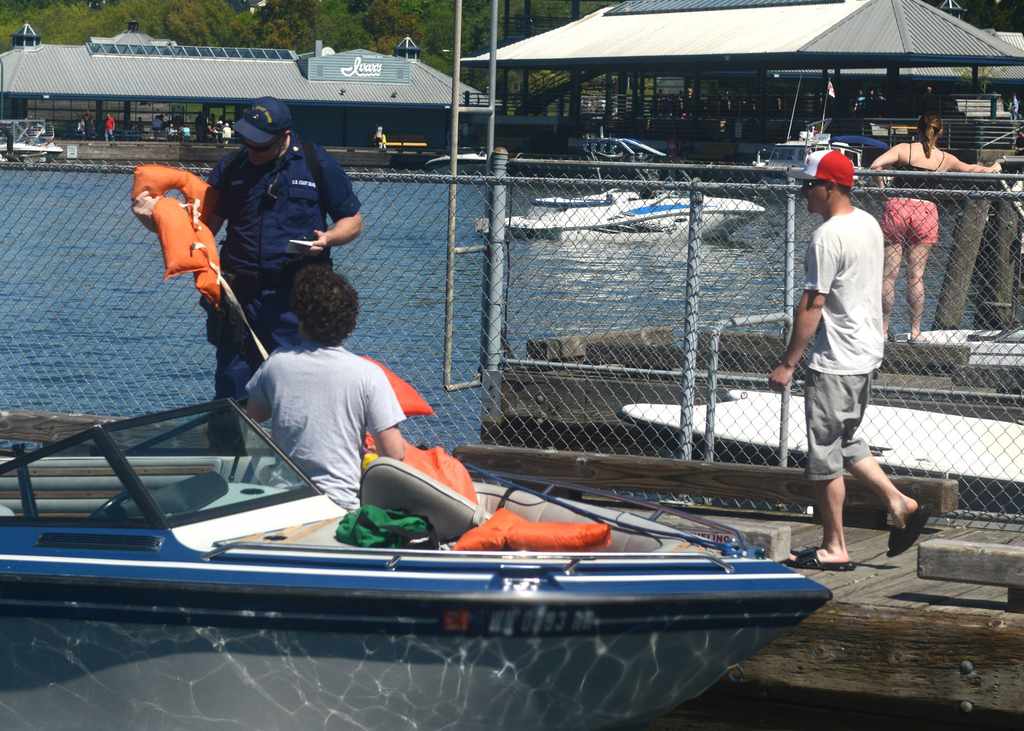 A person in uniform's handing life jackets to two people on a docked boat near a marina, with water and other boats behind them. No visible fish or species like Bass or Trout are present.