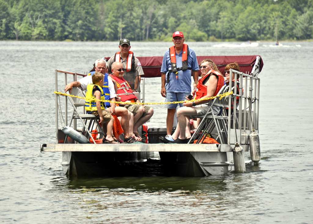 A group of people wearing life jackets are sitting and standing on a pontoon boat that's floating on a lake, with trees in the background.