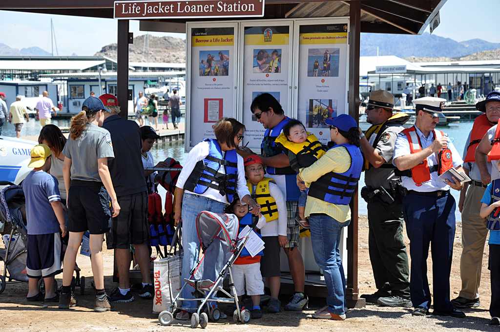 People, including kids and park rangers, gather at a life jacket loaner station by the water; several are wearing life jackets and standing in line.