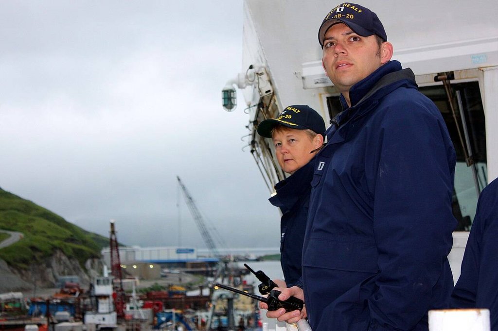 Two people in navy blue uniforms and hats are standing on a ship—one's holding a radio—looking out over a busy industrial port filled with cranes and equipment in the background.