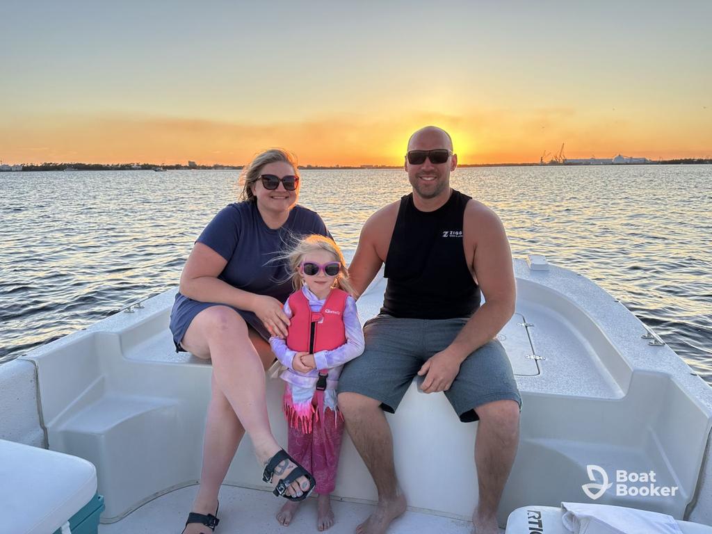 Three people, two adults and one child, are sitting on a boat at sunset and they're all wearing sunglasses. The water and shoreline are in the background. Boat Booker logo's in the corner.