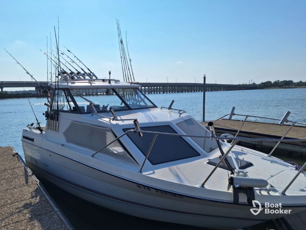 A white motorboat with fishing rods mounted up top's docked at a pier, while a bridge and calm water are in the background.
