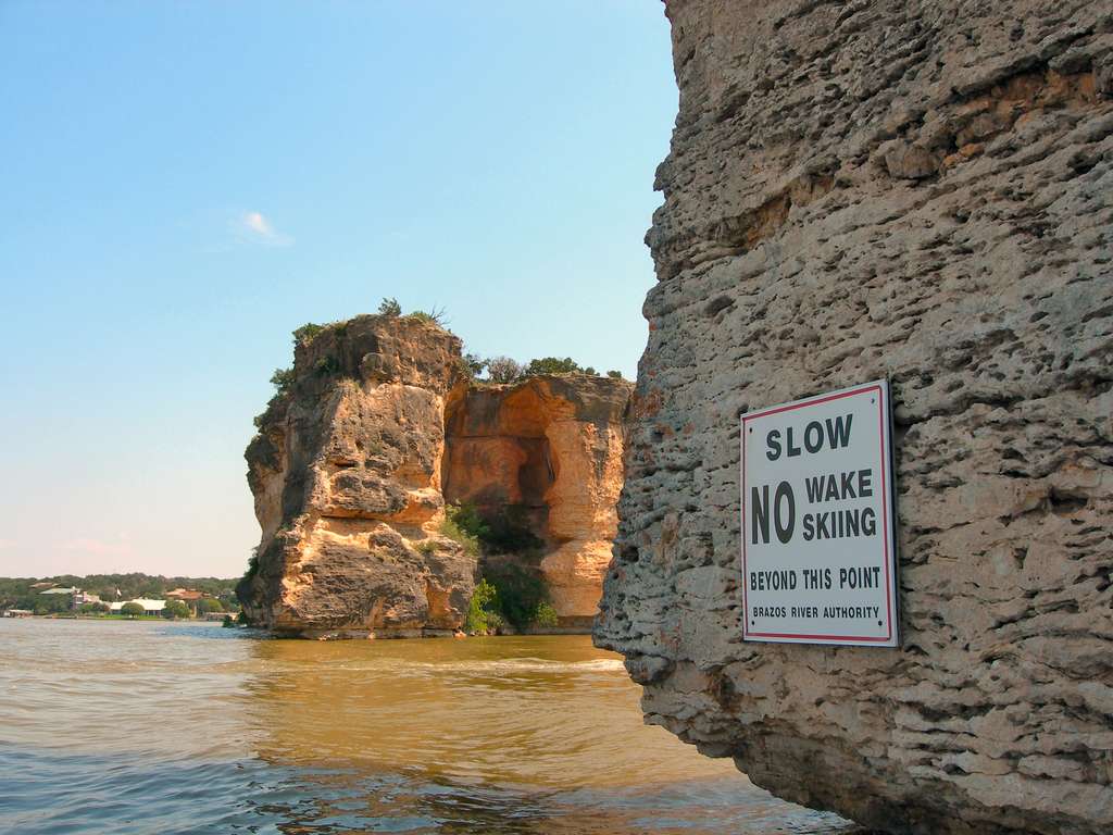 There's a sign on a rocky cliff by the water that says, "Slow No Wake Skiing Beyond This Point," with other cliffs and calm water in the background.