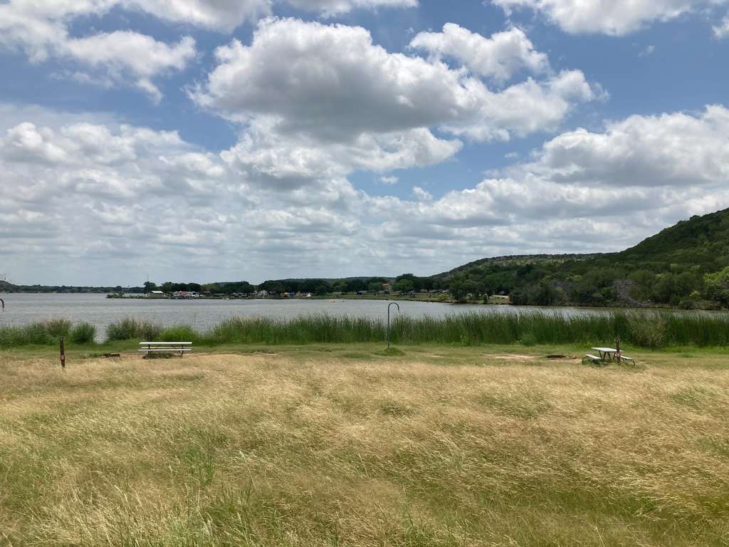A grassy field with two empty picnic tables overlooks a lake bordered by reeds, with hills and a cloudy sky in the background.