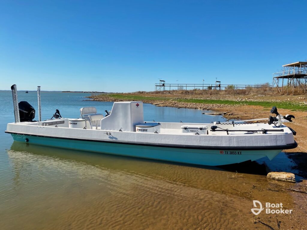 A white center console fishing boat's docked at the shore of a calm lake on a clear day, with grassy banks and several structures in the background.