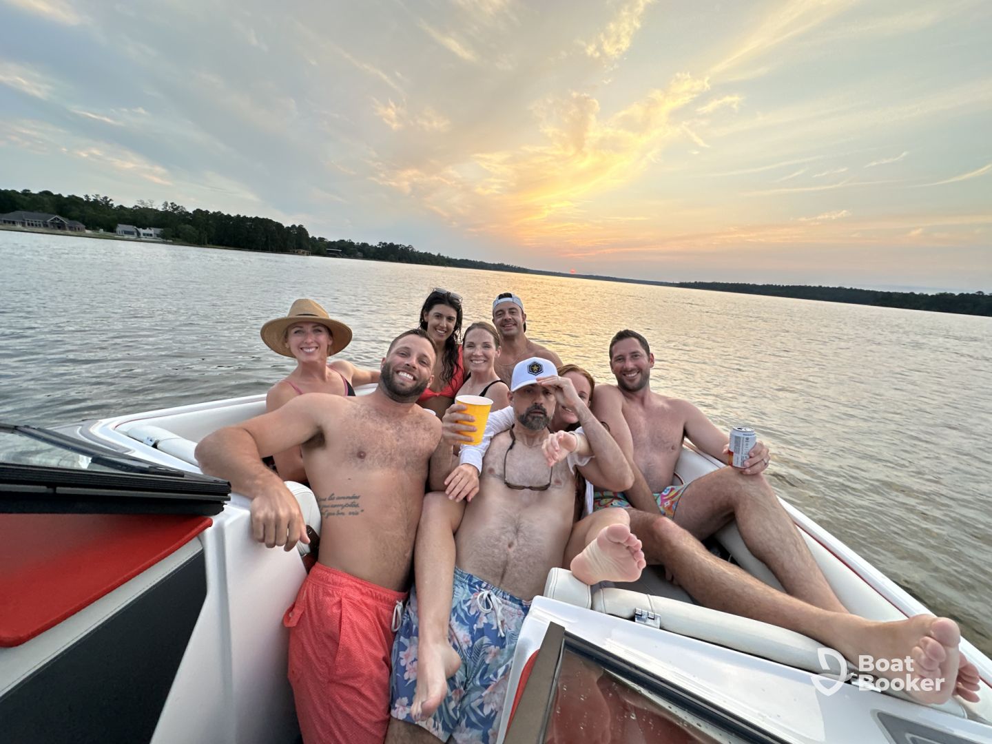 A group of seven adults are relaxing and posing for a photo on a boat at sunset, holding drinks and smiling. The calm water and partly cloudy sky are in the background.