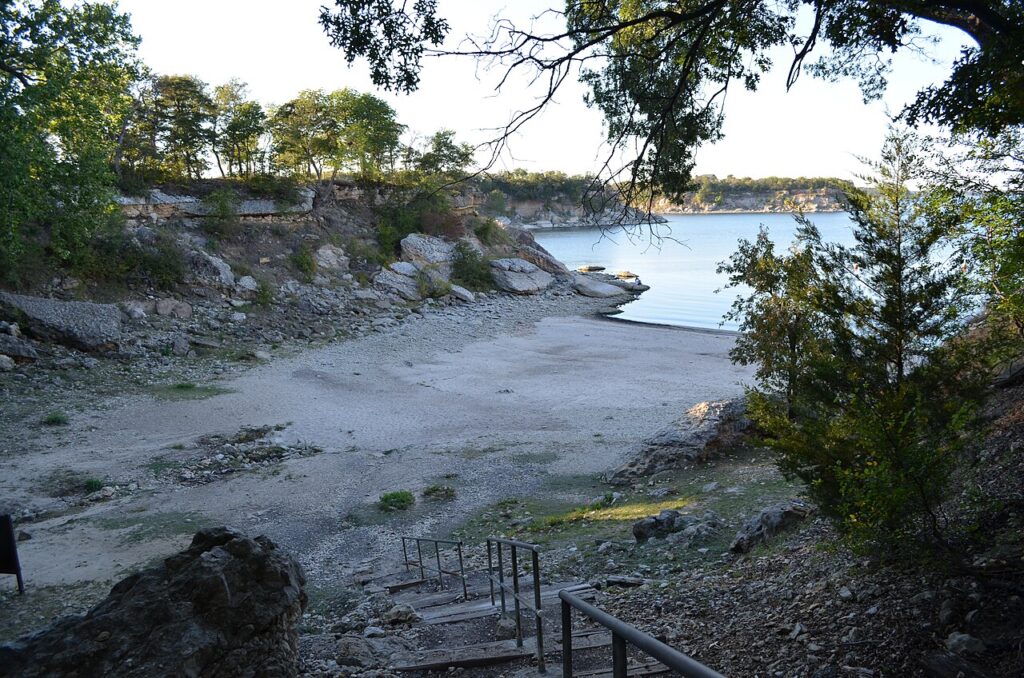 There’s a rocky shoreline and sandy area by a calm body of water, with trees in the background and a metal-railed stairway leading down to the shore.