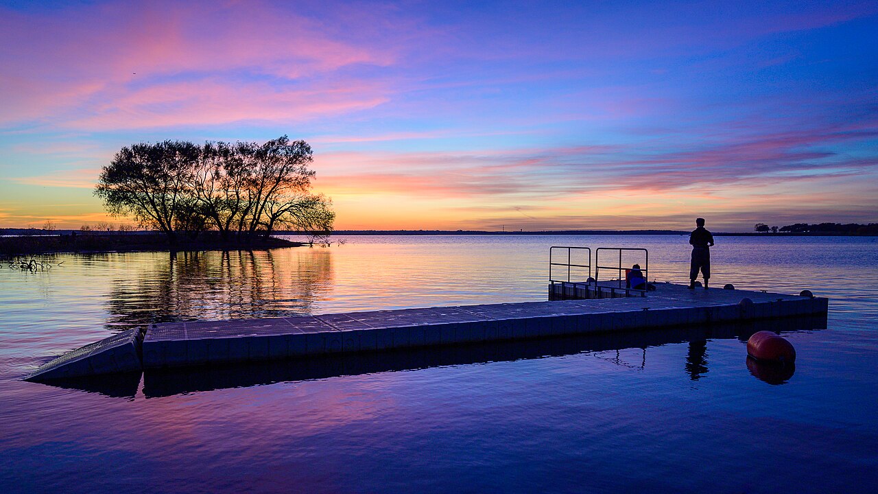 Two people stand on a dock overlooking a calm lake at sunset, with a colorful sky and a small cluster of trees in the water nearby.