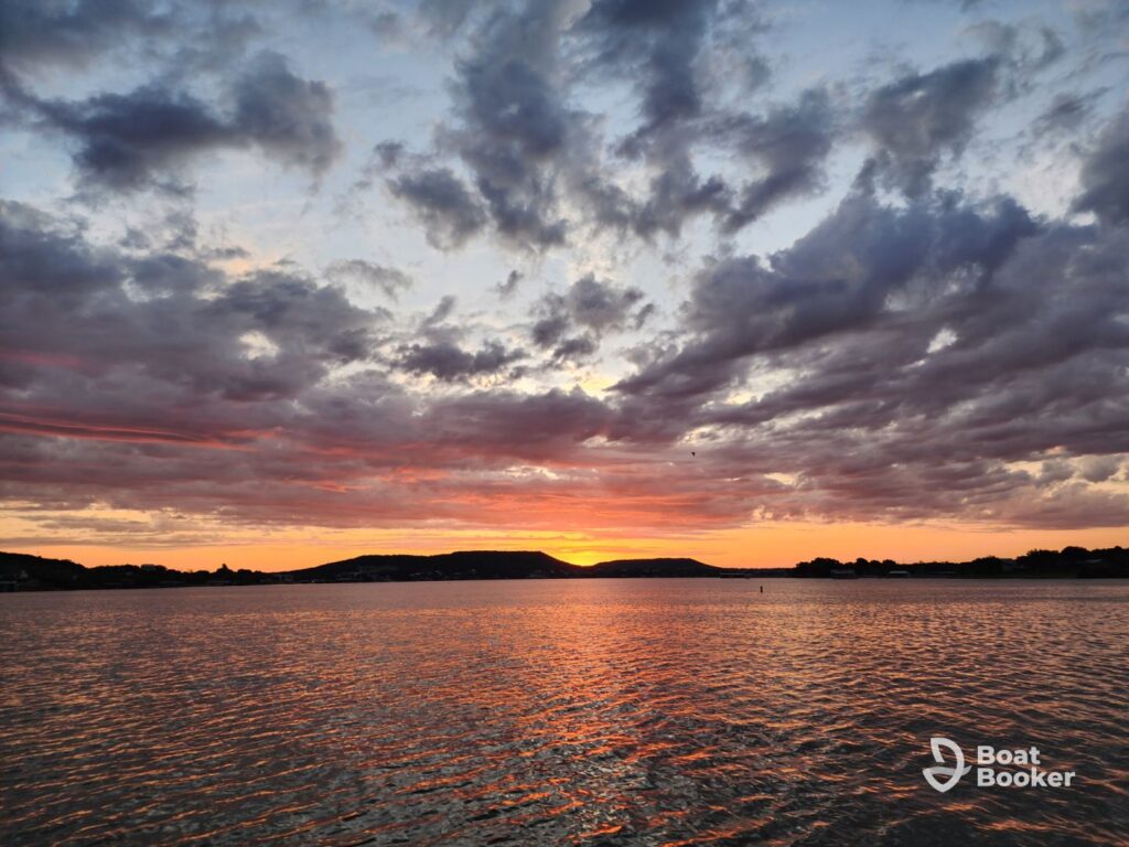 Cloudy sunset over a calm lake with distant hills on the horizon. The Boat Booker logo's in the lower right corner.