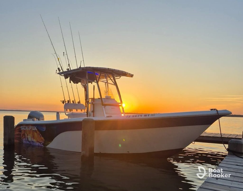 A fishing boat with several fishing rods is docked at a pier during sunset, and the sun's shining behind the boat.