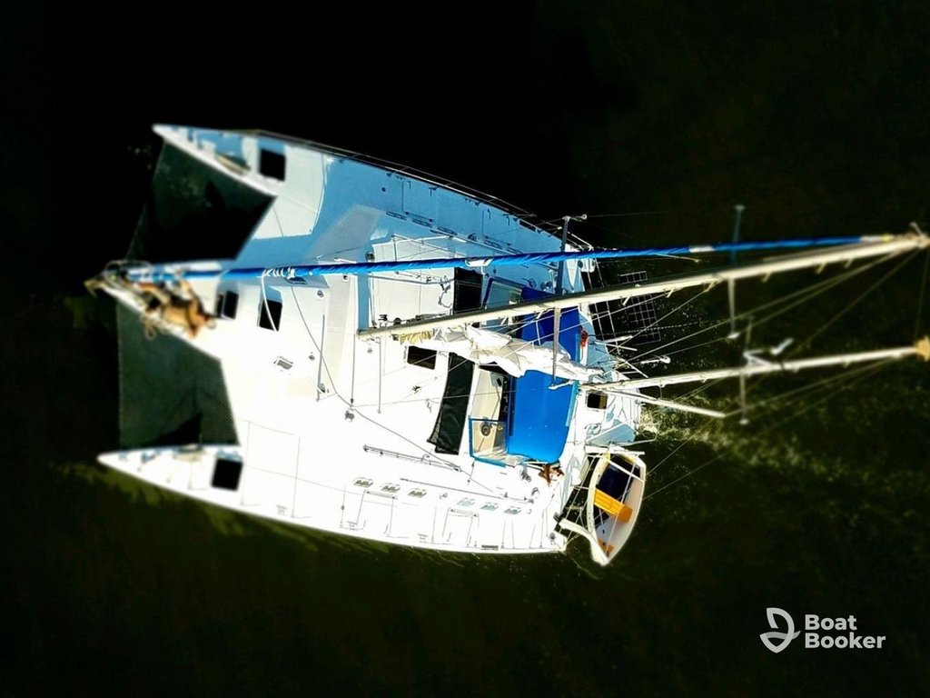 You’re looking down at a white catamaran with a blue canopy and rigging, sailing on dark water; Boat Booker logo’s in the bottom right corner.