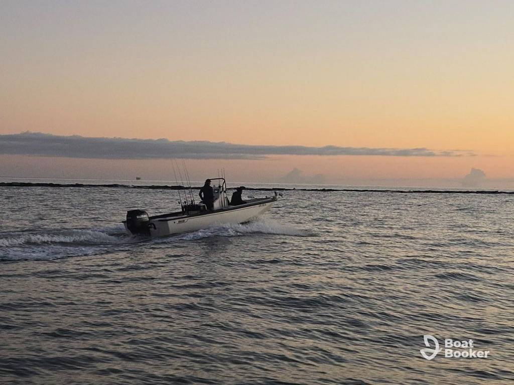 A small motorboat with two people on board moves across calm water at sunset, leaving a gentle wake. The sky's orange and purple, and the “Boat Booker” logo shows in the lower right corner.