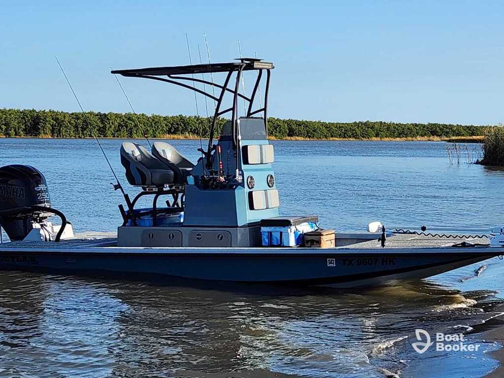 A small blue motorboat with a T-top roof and seating's floating on calm water near a shoreline with trees in the background.