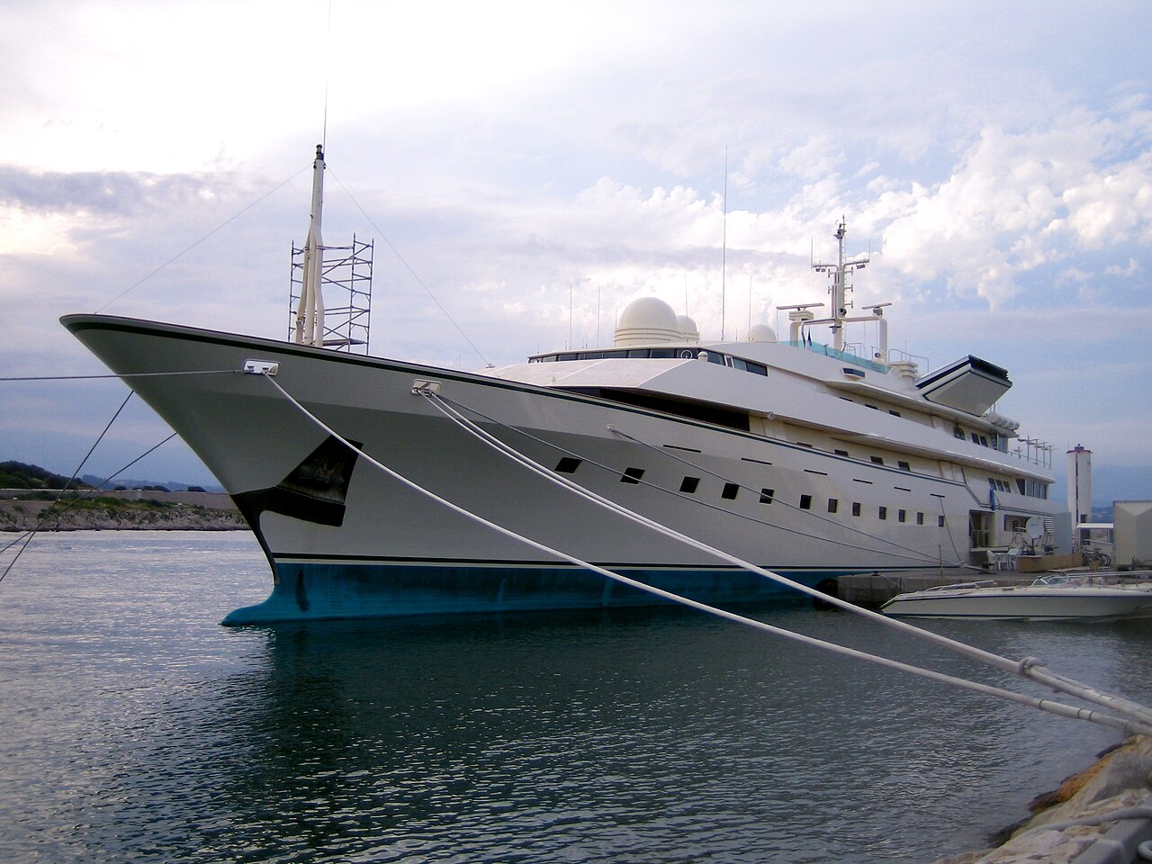 A large white luxury yacht's docked at a marina, secured with ropes, under a partly cloudy sky.
