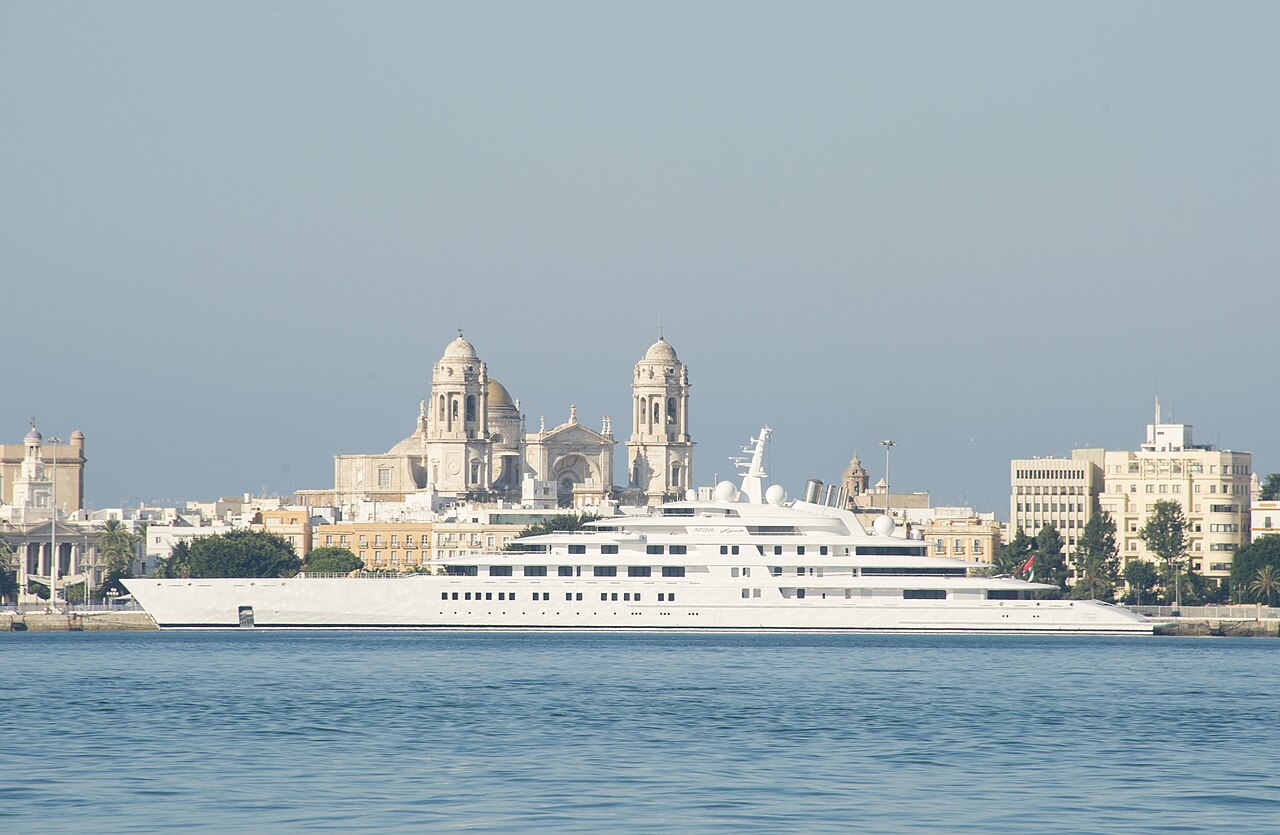 A large white yacht, the Azzam, is anchored in a harbor, with historic buildings and a Cathedral visible in the background under a clear sky.