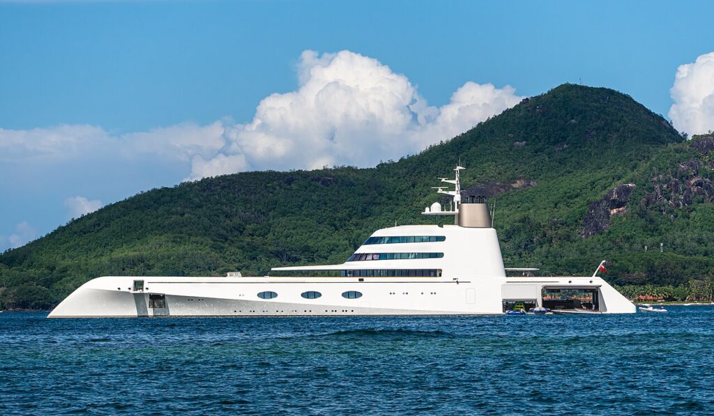 A large, modern white yacht's anchored near a lush green hillside, with blue sky and clouds in the background.