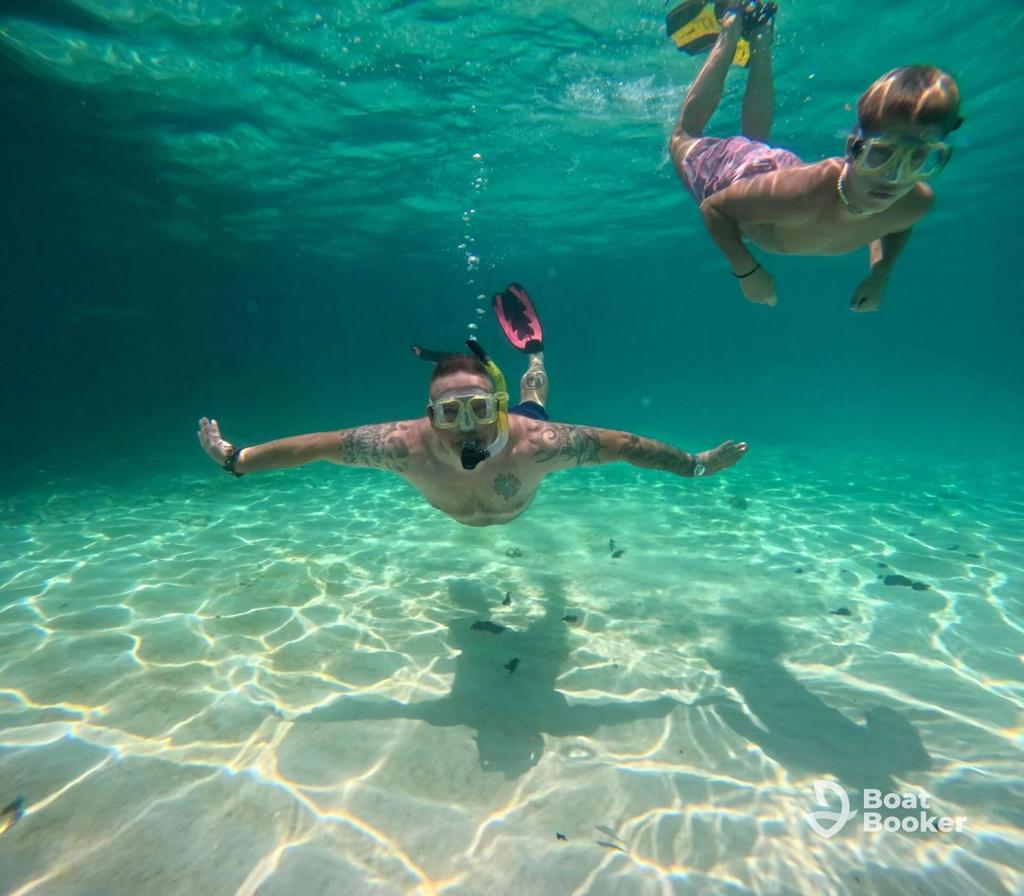 Two people wearing snorkel masks and fins are swimming underwater in clear, shallow water, where you can see sunlight patterns on the sandy bottom. No visible fish species are present.