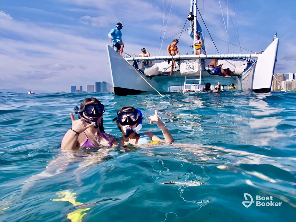 Two people wear snorkel masks and fins as they pose in the ocean near a catamaran with several people aboard. City buildings are visible in the background.