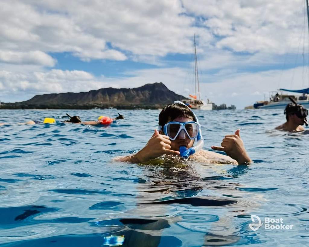 Someone in snorkeling gear's giving a shaka hand sign in the ocean, with boats, other swimmers, and a mountain behind them. No FISH species or animals are visible.