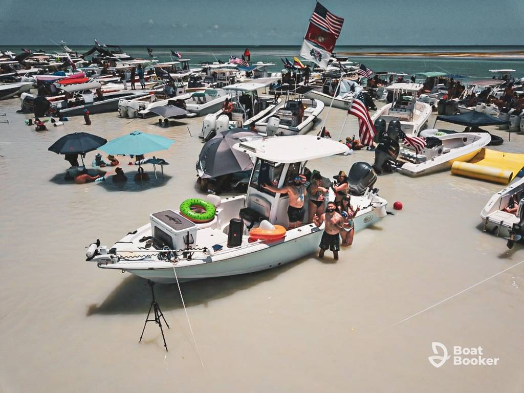 A crowd of people gather on and around several boats in shallow water, with umbrellas and flags visible. The weather's clear and sunny. The "Boat Booker" logo appears in the corner.