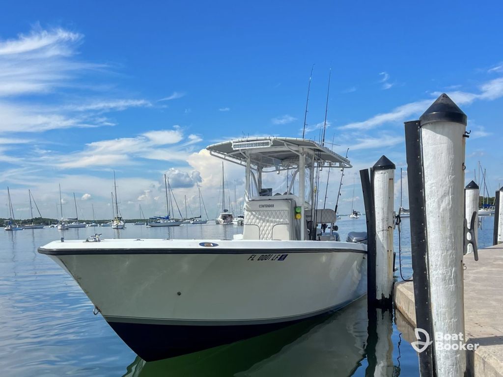 There's a white center console boat docked at a marina beside wooden pilings, with sailboats anchored in the background under a blue sky.