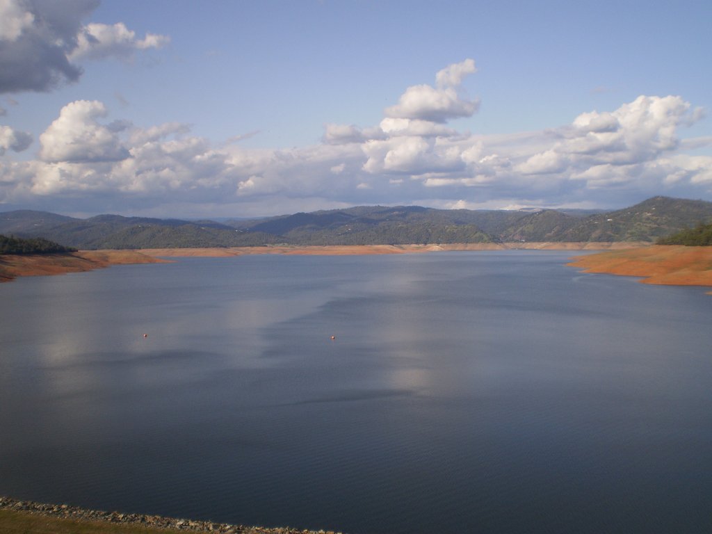 A large reservoir's surrounded by dry, brown banks and distant green hills under a partly cloudy sky.