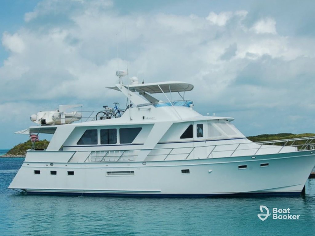 There's a white motor yacht with large windows anchored in calm blue water by a green shoreline, under a cloudy sky. The Boat Booker logo's visible in the corner.