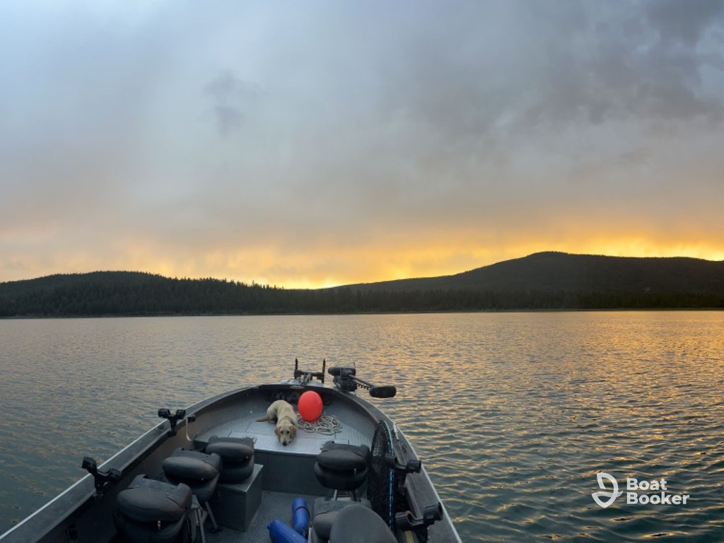 A small boat with empty seats and a red balloon floats on a calm lake at sunset, with tree-covered hills in the background. The "Boat Booker" logo's in the bottom right corner.