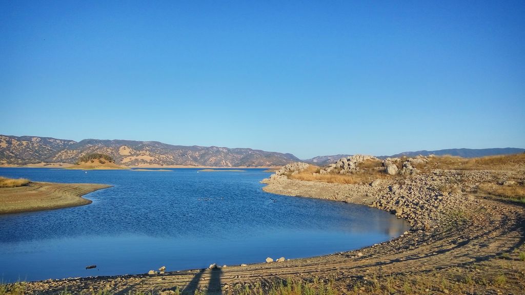 A calm lake with rocky and grassy shores under a clear blue sky, surrounded by distant hills and mountains. No visible fish like Trout or Bass in the water, just peaceful natural scenery.