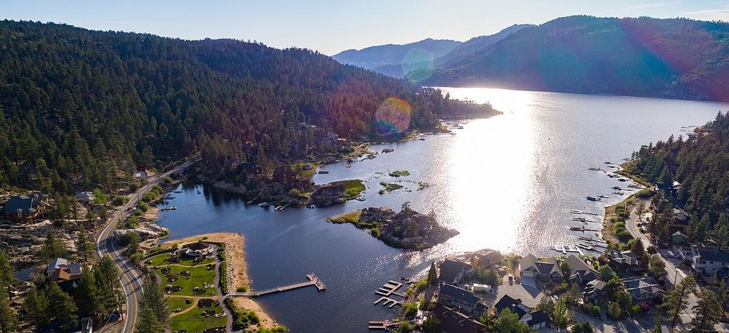 You’re looking down on a lake encircled by forested hills, homes, and docks, with a sandy shoreline. The clear sky’s bright as sunlight glints off the water—no Trout or Bass visible from above.