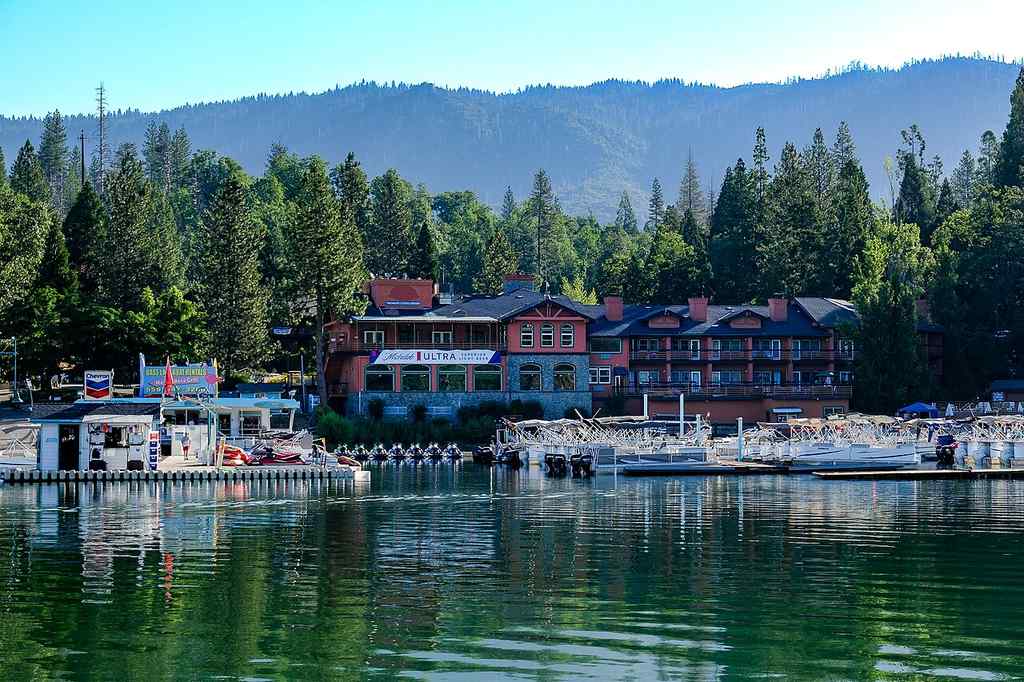 A marina with docked boats sits in front of a lakeside lodge surrounded by Pine trees and mountains in the background.