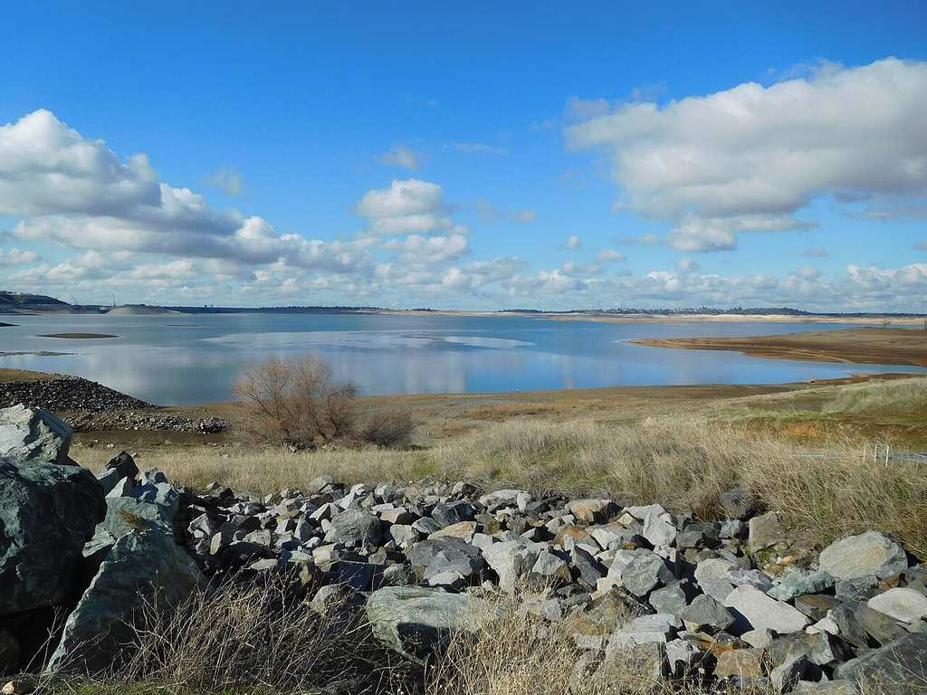 There’s a calm lake with rocky shorelines and dry grass beneath a blue sky that’s dotted with scattered clouds.