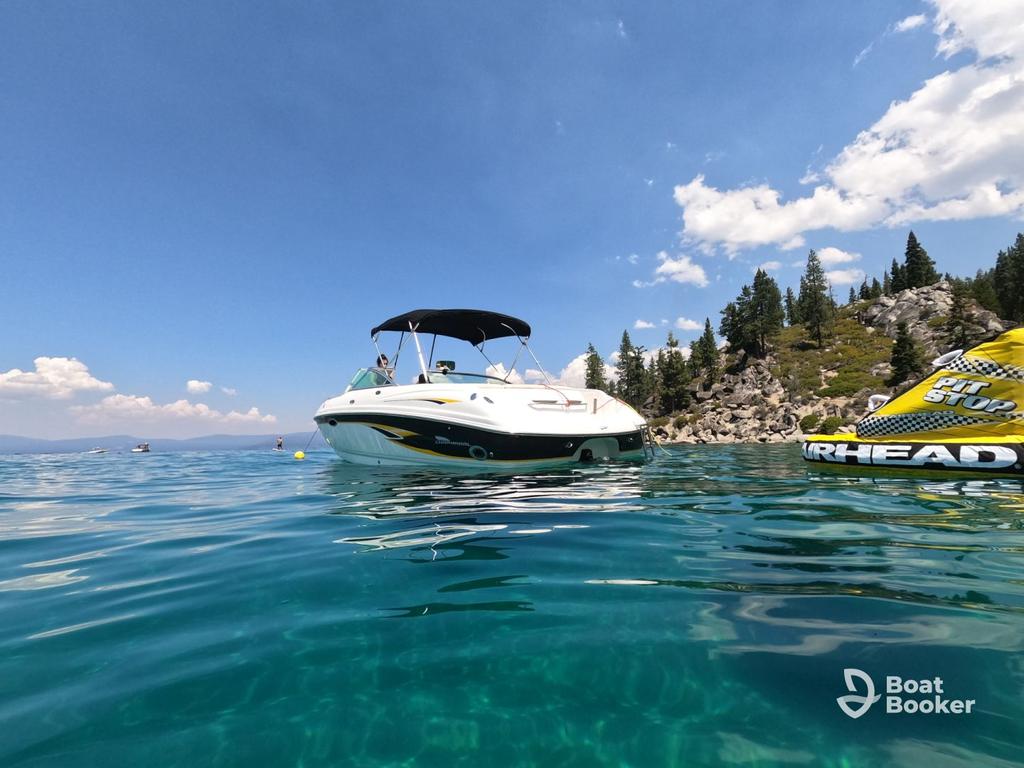 A white motorboat with a black canopy floats on clear blue water near a rocky, tree-covered shore under a partly cloudy sky. An inflatable labeled "AIRHEAD" is nearby.