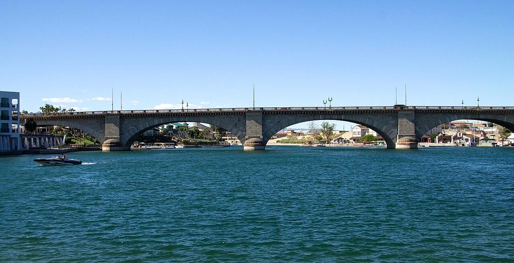 A stone arch bridge spans a wide blue river under a clear sky, with a boat gliding on the water and buildings in the background.