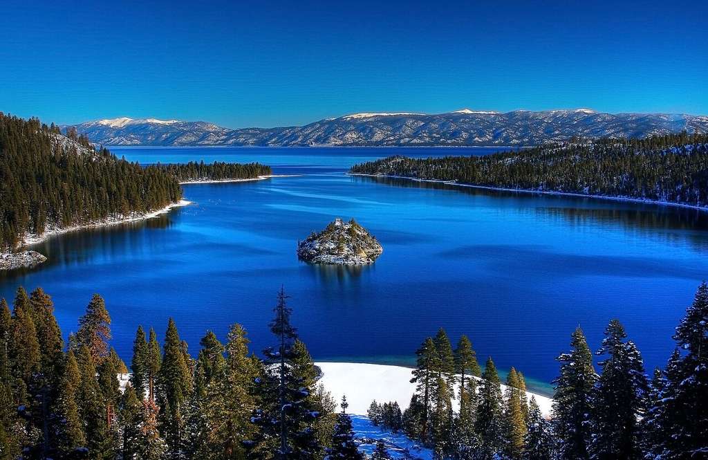 You’ll see a clear blue lake ringed by pine-covered hills and mountains, with a small island at its center and snow on the landscape.
