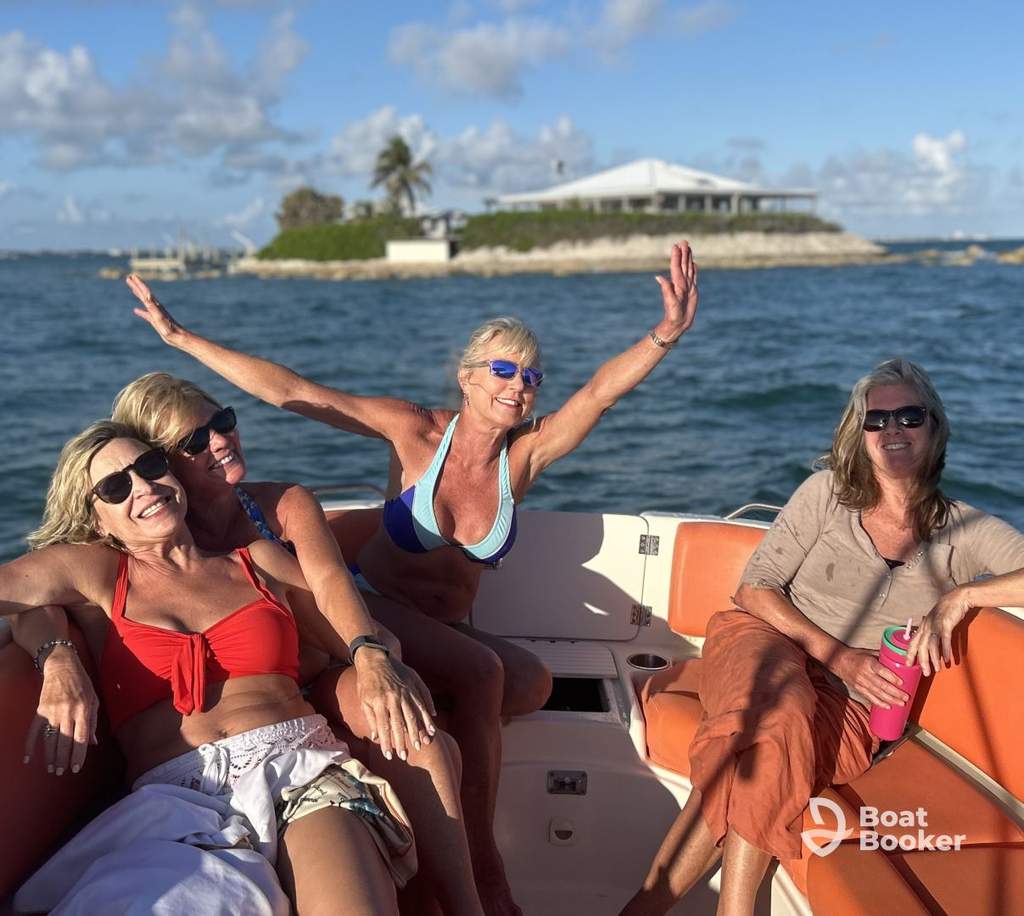 Four women are smiling and posing on a boat in sunny weather near an island house. The "Boat Booker" logo's visible—it's perfect for travelers with a Florida boating license.