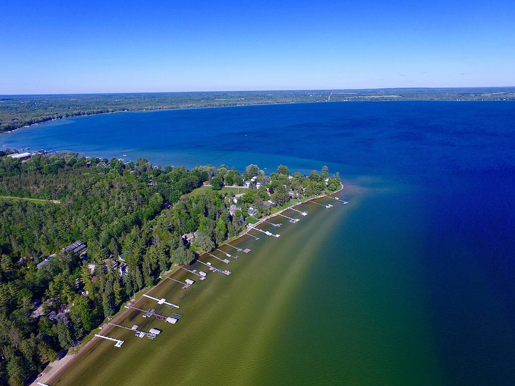You're seeing an aerial view of a lakeshore with seven docks stretching into the water, surrounded by green trees and clear blue skies.