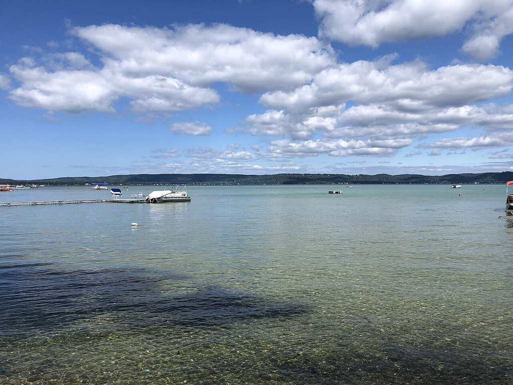 There's a clear, shallow lake with a dock and boat under a partly cloudy sky. You can see the distant tree-lined shore across the water.