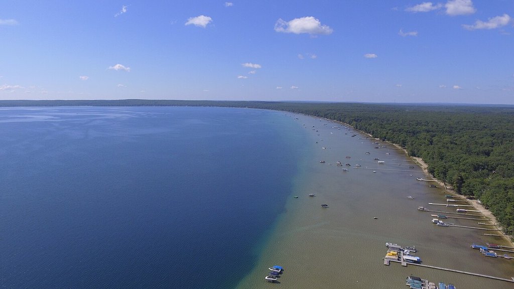 You’re looking down on a big lake with clear blue water, forested shoreline, docks, and boats under a mostly clear sky with just a few small clouds.
