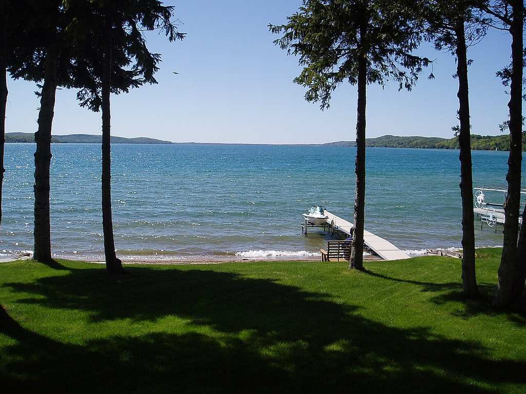 You’ll see a blue lake bordered by trees and green grass, with a wooden dock stretching into the water beneath a clear sky.