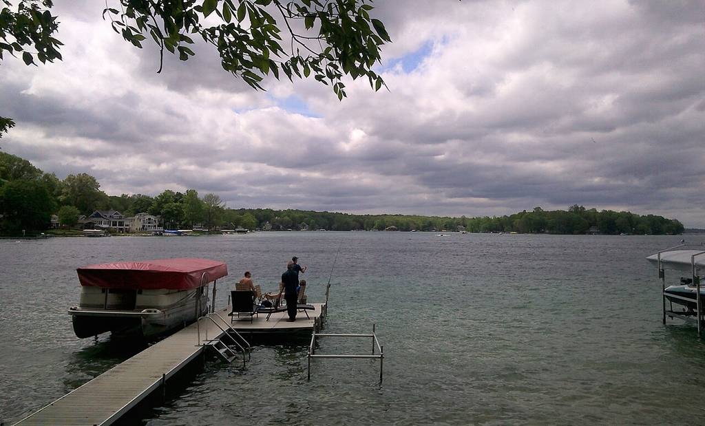 Three people sit and stand on a dock by a covered boat, looking out over the lake under cloudy skies, with trees and houses in the distance.