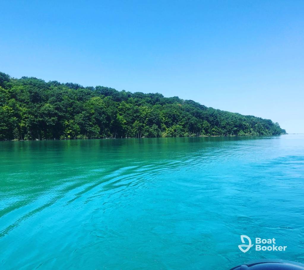 There's a calm turquoise lake with gentle ripples, surrounded by dense Green Forest under a clear blue sky. The "Boat Booker" logo's in the bottom right corner.