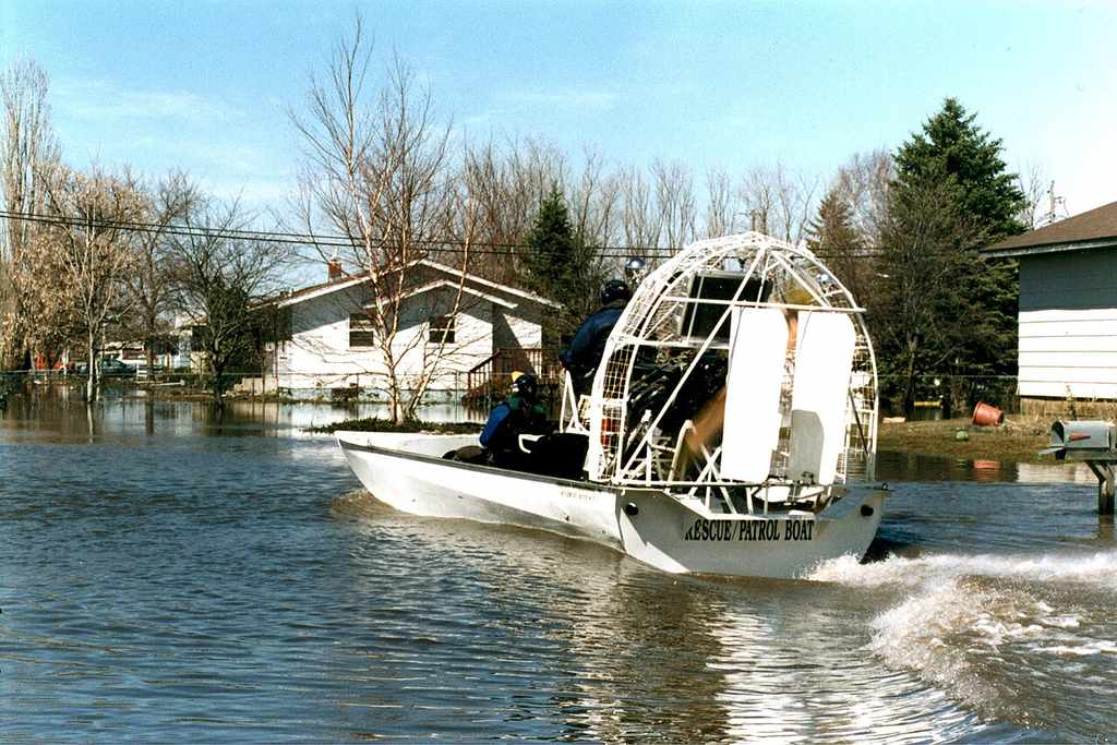 An airboat marked "Rescue Patrol Boat" glides through a flooded neighborhood with two people aboard, showing what airboats can do in emergencies.