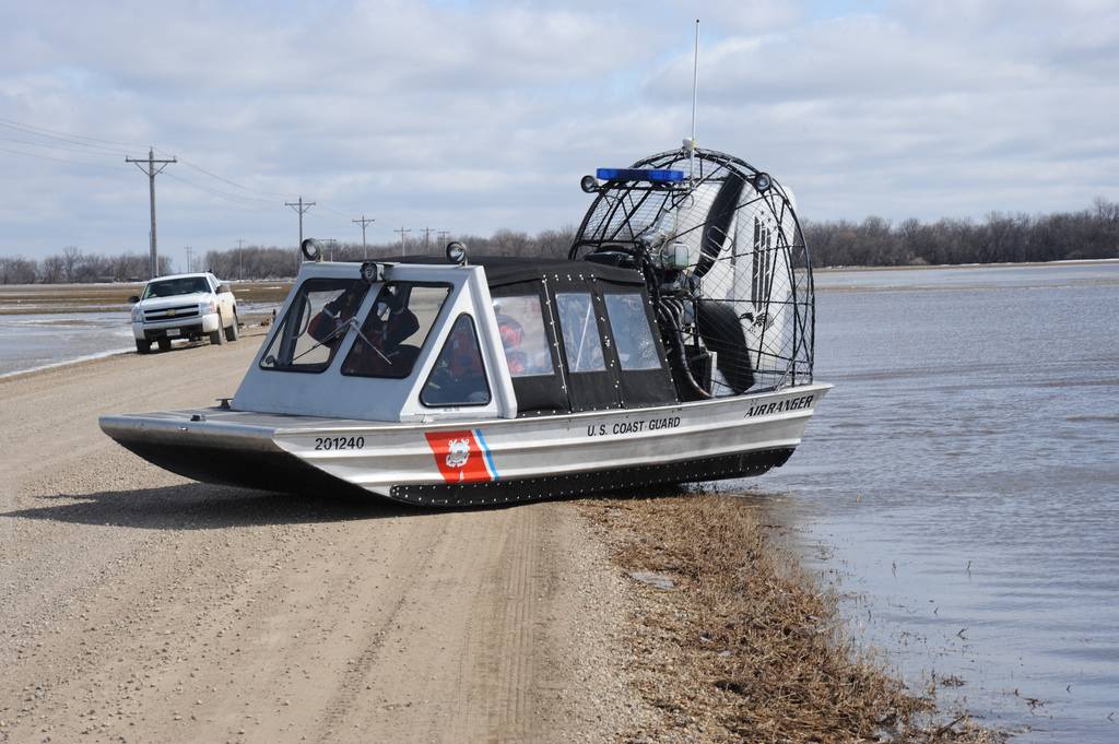 A U.S. Coast Guard airboat—what's an airboat, you ask?—sits partly on a dirt road and partly on water next to a white pickup truck.