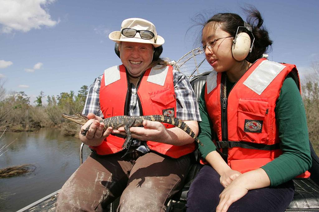 Two people in red life vests and ear protection, maybe after asking "what's an airboat," sit outside; one holds a small Alligator by water and trees under a blue sky.