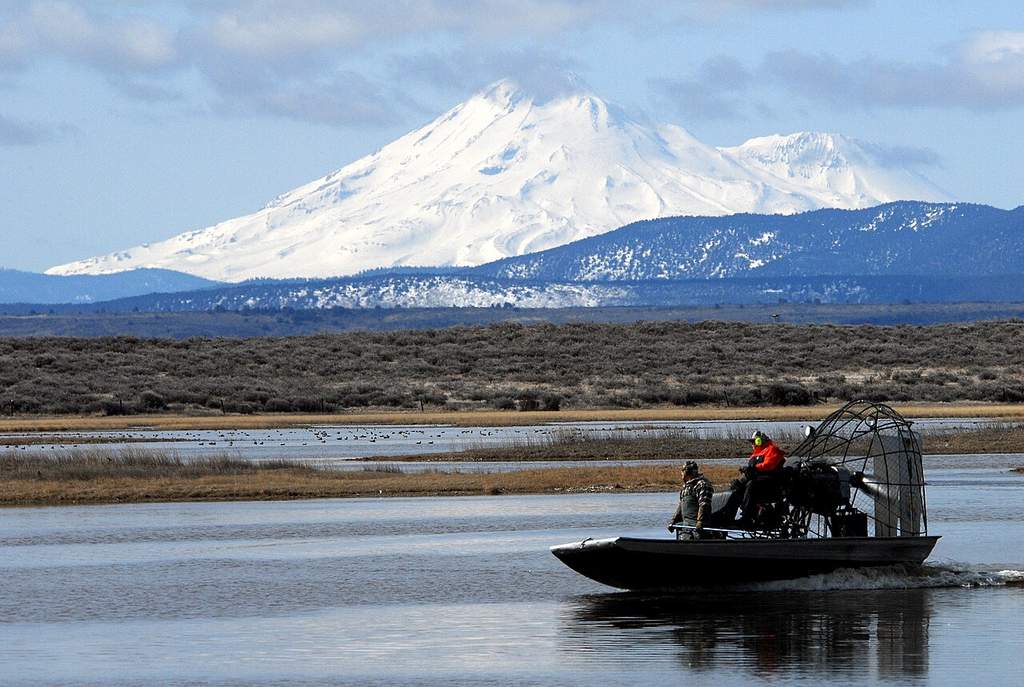 An airboat's gliding across a lake with passengers, set by rugged terrain and a snow-capped mountain under cloudy skies.