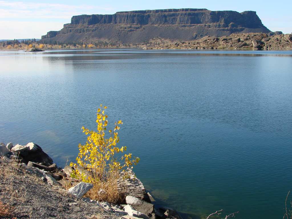 A calm lake—one of Washington State's best for boating—has a rocky shore and yellow-leaved shrub. A flat-topped mesa rises in the background under a clear sky.
