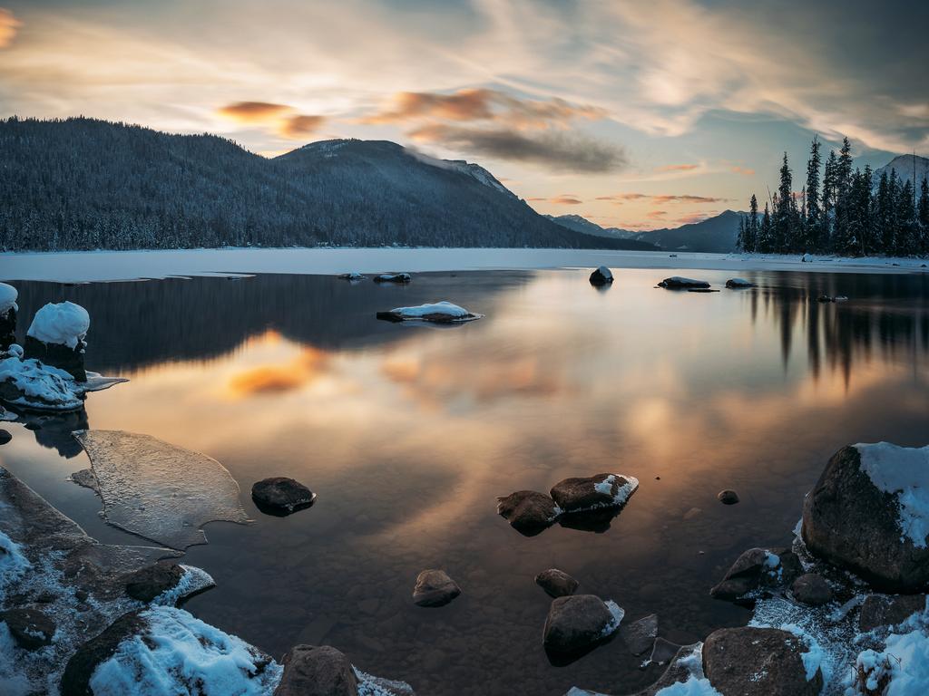 A snowy lakeshore with scattered rocks and ice reflects a colorful sunset sky—it's one of the best boating lakes in Washington, framed by mountains and evergreen trees.